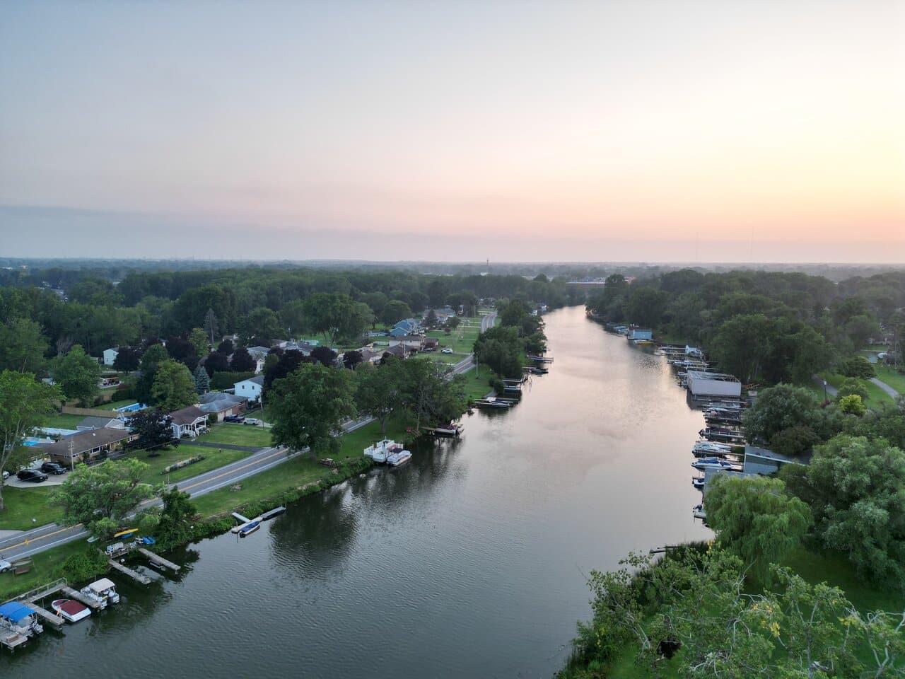 Aerial view of a serene river flowing through a residential area with houses and docked boats on either side during dusk, showcasing a gradient sky from blue to orange. buffalo aerial videography and drone services